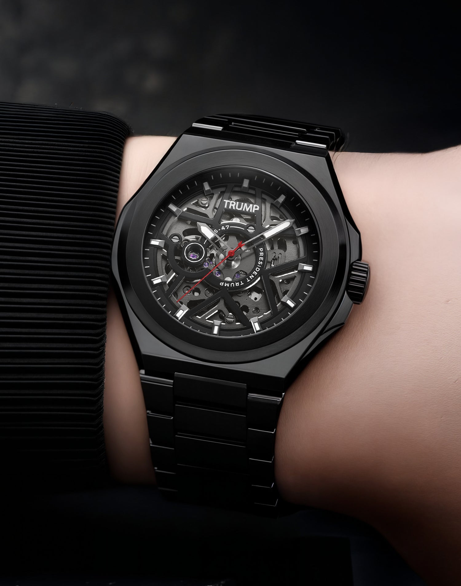 Black skeleton dial Trump wristwatch with red second hand and metal link bracelet on wrist.