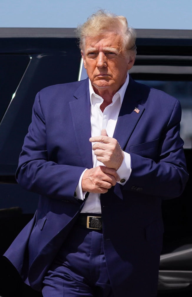 Man in navy blue suit adjusting cuff, standing beside black vehicle under bright daylight.
